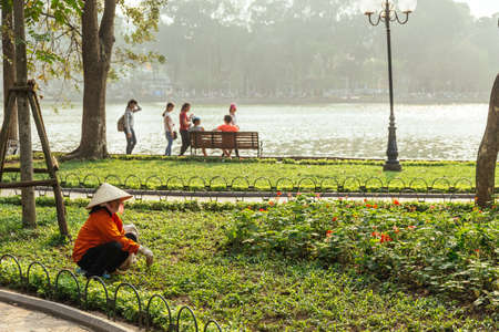 Red cloth woman gardener wearing Vietnamese conical hat cut grass in yard of outdoor park near  Hoan Kiem Lake in Hanoi, Vietnam.のeditorial素材