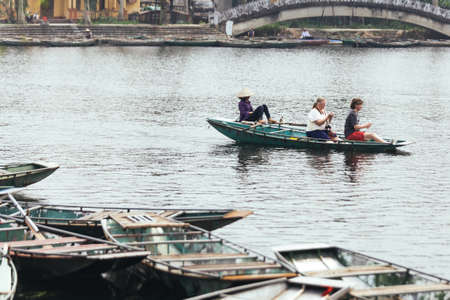 Woman wearing purple long arm t-shirt and conical hat rowing boat by her feet with tourists on the boat on the river at Trang An Grottoes in Ninh Binh, Vietnam.のeditorial素材