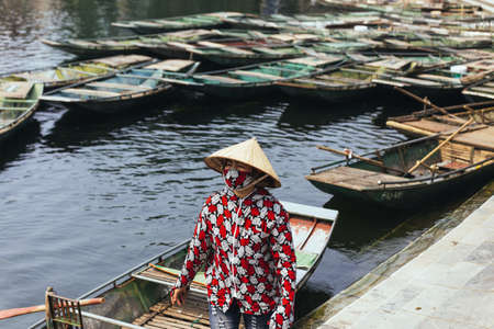 Rowing boat woman wearing red and white colors shirt, conical hat and mouth mask standing with empty boats over the river in background in Ninh Binh, Vietnam.のeditorial素材