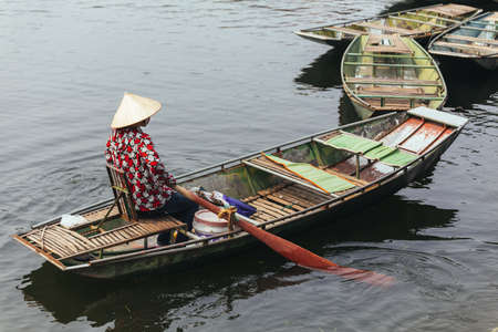 Rowing boat woman wearing red and white colors shirt, conical hat and mouth mask sitting in a boat with paddles over the river in background in Ninh Binh, Vietnam.のeditorial素材