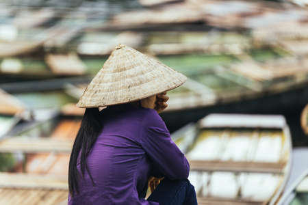 Vietnamese woman wearing purple long arm t-shirt conical hat siting and waiting on the land for tourist at Trang An Grottoes in Ninh Binh, Vietnam.の写真素材