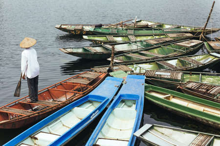 Man wearing white shirt, conical hat with his paddle stand on red empty rowing boat with many boats stop over the river at Trang An Grottoes in Ninh Binh, Vietnam.の写真素材