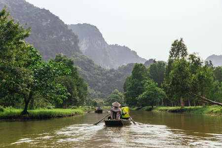 Rowing woman row a boat with tourist on the river along the way with plants and trees and mountain in the background at Trang An Grottoes in Ninh Binh, Vietnam.のeditorial素材