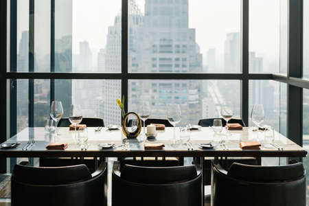 Six people dinner table with plates, knives, forks, wine glasses, glasses and napkins on marble table top with Bangkok cityscape in background.のeditorial素材