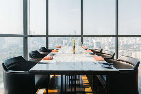 Six people dinner table with plates, knives, forks, wine glasses, glasses and napkins on marble table top with Bangkok cityscape in background.のeditorial素材