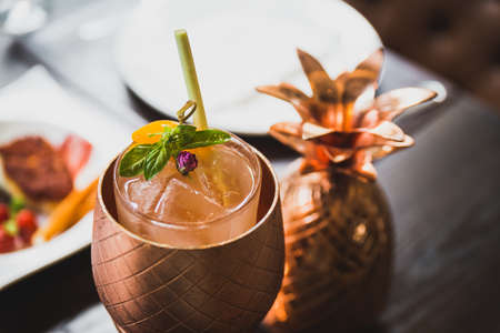 Cocktail served inside copper pineapple cup with dishes in the background.