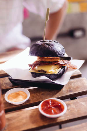 Beef burger with a black bun with cheese, fried bacon and onion rings served with ketchup and mayonnaise.の写真素材