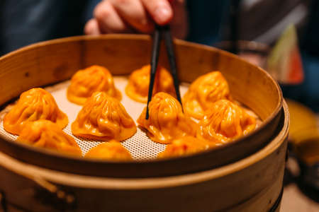 A hand is pinching Orange color Foie Gras Xiao Long Bao (Chinese Soup Dumpling) with chopsticks from bamboo streamer.の写真素材