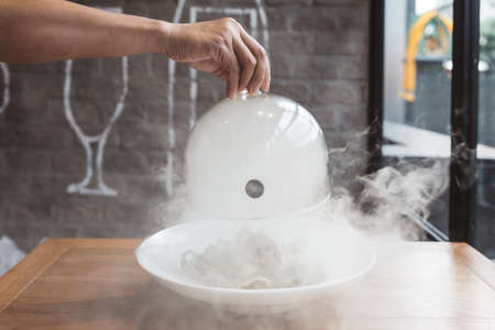 Male hand lifts to up glass cloche from a plate with hot food and moving smoke at the restaurant - Smoked food time! Close Up.の写真素材