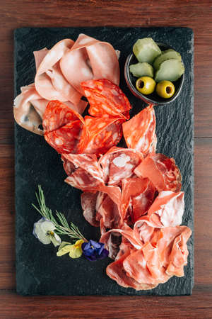 Cold cuts on stone plate with prosciutto, bacon, salami and sausages decorated wth flowers on wooden...