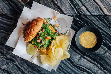 Top view of Roasted chicken salad burger served with chips and mustard sauce on marble top table.