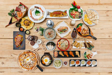 Flatlay of International Foods on wooden table: Sushi, Sashimi, Nachos, Spaghetti, Naan, Sandwich, Noodle, Steak, Ramen. Yakitori, Pizza, Bibimbap, Fried Chicken Wings, Wonton Soup and Brownie Frappe.の写真素材