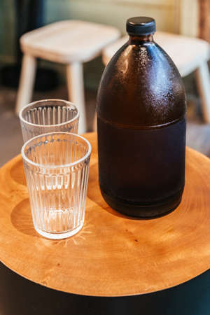 Cold espresso coffee inside black bottle on wooden table with empty drinking glasses.