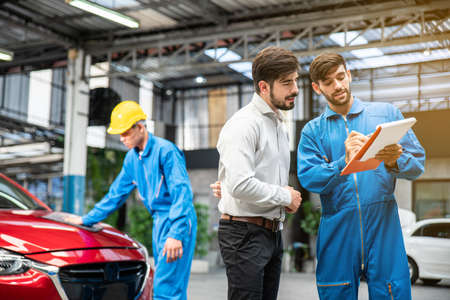 Mechanic show the car checking list for customer with blur his assistant checking red car in the background. Auto car repair service center. Professional service.の写真素材