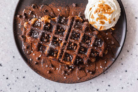 Crop image flat lay of yummy homemade Belgian waffle topping with dark chocolate syrup, cocoa powder and chocolate chips, served with whipped cream in brown color plate. Delicacy and stylish cuisine.の写真素材