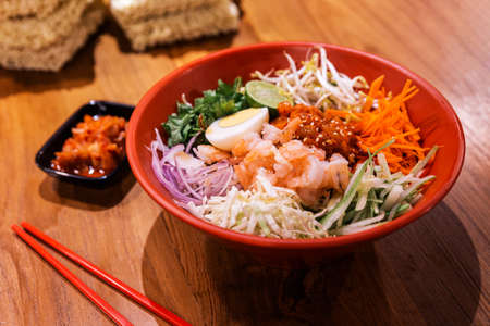 Shrimp Bibimbap Noodles, Korean noodles mixed with radish, sprout, carrot, cabbage, cucumber,  boil egg and Gochujang in red bowl served with Kimchi and red chopsticks. Perfect mouth watering cuisine.の写真素材