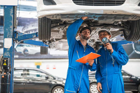 Mechanic in blue work wear uniform inspects the car bottom with his assistant. Automobile repairing service, Professional occupation teamwork. Vehicle maintenance.の写真素材