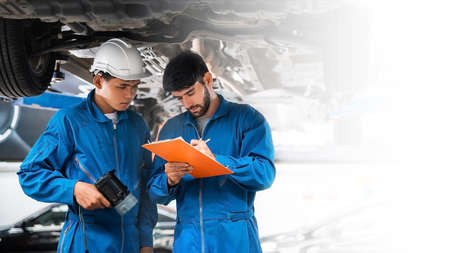 Mechanic in blue workwear uniform checks the vehicle maintenance checklist with his assistant under...