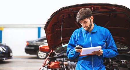 Mechanic in blue workwear uniform checks the vehicle maintenance checklist in a smartphone while...