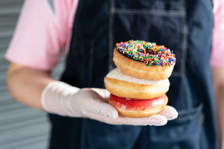 Stacking chocolate frosted donut with sprinkles, sugar-glazed frosted and Bavarian donut on woman hand that wearing latex gloves. Playful and joyful tasty sugary comfort food for customers.の写真素材
