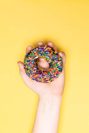 Hand holding chocolate frosted donut with sprinkles, sugar-glazed frosted on yellow background. Playful and joyful tasty sugary comfort food for customers with copy space. Donut lovers.の写真素材