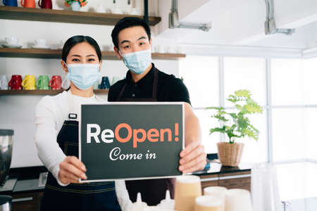 Young baristas couple wearing mask opening their own cafe, making announcements with a sign on a board "ReOpen! Come In ". Enthusiastic, startup business while coronavirus pandemic concept.の写真素材