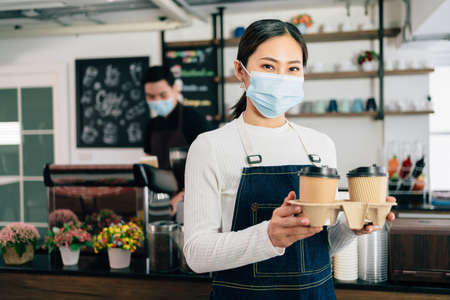 Young female barista wearing a face mask holding coffee in takeaway paper disposable cups in the coffee shop. Coffee making classes for entrepreneurs to start a small business. Women leading.の写真素材