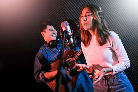 Pretty Asian female singer recording songs by using a studio microphone and pop shield on mic with male playing guitar in blue and red light. Performance and show in the music business. Duet session.の写真素材
