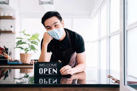 Young male barista cafe owner wearing a face mask, standing inside the coffee counter and holding the "OPEN" announcement board. People's lifestyles during Covid-19 pandemic.の写真素材