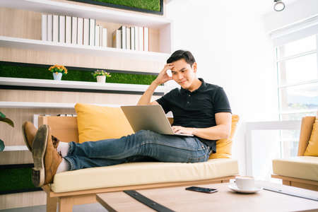 Asian male in casual feels stress and concerned with laptop while working and sitting on the sofa....