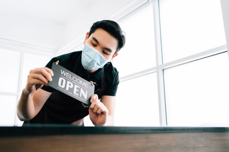 Young male barista cafe owner wearing a face mask, standing inside the coffee counter and holding the "OPEN" announcement board. People's lifestyles during Covid-19 pandemic.の写真素材