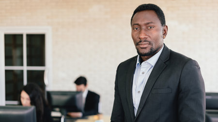 Confident African American businessman standing in office with blur employees working on their desk in background. African American businessman portrait.の写真素材