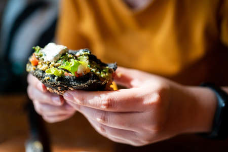 Hands cuts Broccoli quinoa charcoal burger topped with guacamole, mango salsa and fresh salad by knife and fork, served in a white plate. Creative vegan meal for vegetarians.の写真素材