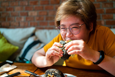 Asian woman bites Broccoli quinoa charcoal burger topped with guacamole, mango salsa and fresh salad in hands with happy mood. Creative vegan meal for vegetarians.の写真素材