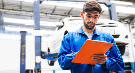 Mechanic in blue work wear uniform checks the vehicle maintenance checklist with blur lifted car in...