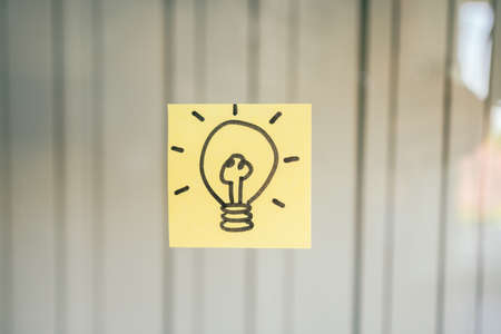 Hand drawing light bulb that draws on a yellow sticky note on glass board with copy space.の写真素材