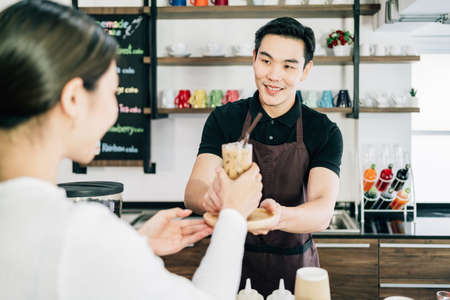 Asian male barista serving iced coffee to a woman in the coffee shop with a smile and happy mood.の写真素材