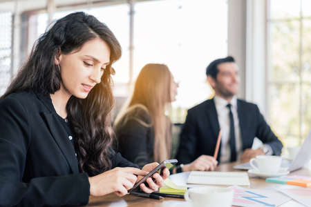 A Caucasian woman manager in a black suit sitting and working on a smartphone, a businesswoman typing on a touch screen of cellphone while meeting in the office with blur colleagues in the background.の写真素材