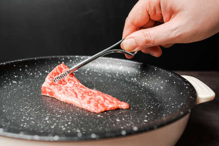 Close up premium rare slices sirloin Wagyu A5 beef with high-marbling texture pick up and grill on marble coated non-stick pan by bbq tongs with hand on black background. Served for Yakiniku.の写真素材