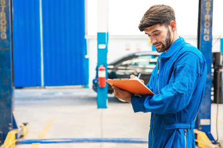 Mechanic in blue work wear uniform checks the vehicle maintenance checklist with blur lifted car in the background. Automobile repairing service, Professional occupation.の写真素材