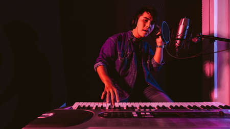 Male playing an electric keyboard while wearing headphones. Recording songs by using a studio microphone and pop shield on the microphone in blue and red light with copy space.の写真素材
