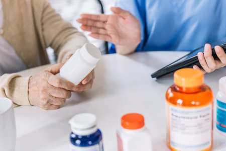Close up hands of a senior woman holding a medical bottle and asking information from the nurse before administering medication. Caregiver visit at home. Home health care and nursing home concept.の写真素材