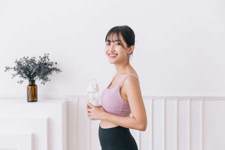 Asian woman with long black hair tied up, happy smiling at the camera wearing sportswear and holding a water bottle standing on a white background. Active lifestyle image with copy space.の写真素材