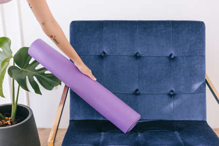 An Asian woman wearing sportswear and yoga pants picks up a rolled-up yoga mat from the chair....