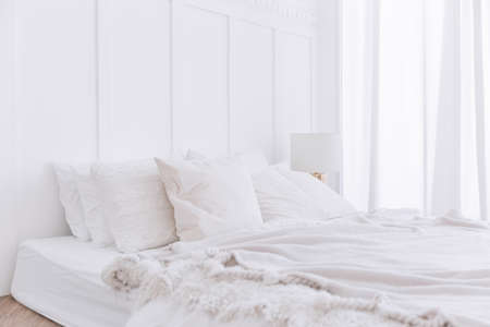Crop image of white bed on the floor with pillows, blankets and lamp. White bedroom decoration with white curtain on the window with natural light.の写真素材