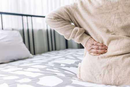 Mature senior woman wearing a sweater holding her pain back in bed. Suffer from lower lumbar...