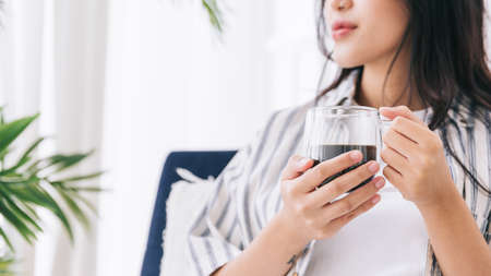 Crop image of woman's hands holding a cup of coffee in the living area with a green plant, Relaxing drinking a cup of hot coffee and enjoying a quiet time and indoor environment at cozy home.の写真素材