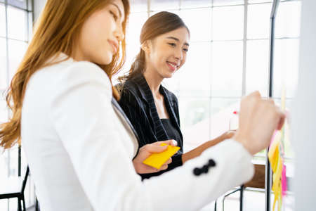 Group of successful Asian businesswomen teamwork. Brainstorm meeting with colorful sticky paper notes on the glass wall for new ideas. Using agile methodology for business in a tech start-up office.の写真素材