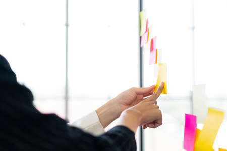 Close up hands of Asian businesswomen. Brainstorm meeting with colorful sticky paper notes on the glass wall for new ideas. Using agile methodology for business in a tech start-up office.の写真素材