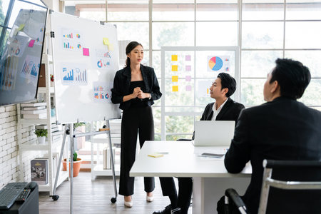 A cheerful and confident Asian businesswoman stands, present bar charts data from a whiteboard to her office colleagues. Asian business women leader role at the meeting.の写真素材